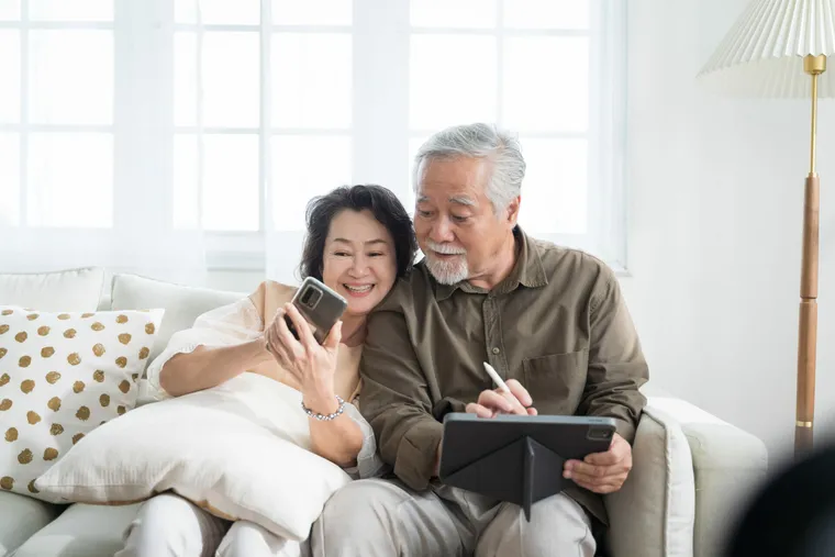 Asian senior couple in living room at home.Wife browsing online on smartphone showing something to her husband while husband is also using a tablet.